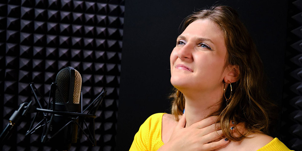 A person standing behind a microphone grimaces while holding their throat.